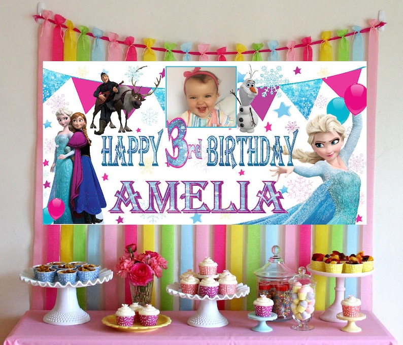 May include: A white banner with a Frozen theme for a 3rd birthday party. The banner features a photo of a young girl, Elsa and Anna from Frozen, Olaf, and a reindeer. The banner says "Happy 3rd Birthday Amelia".