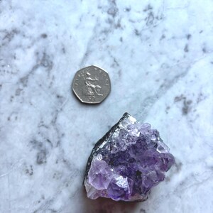 May include: A cluster of amethyst crystals with a deep purple hue, resting on a marble surface. A 50-pence coin is next to the crystal for size comparison. The amethyst has a rough, natural texture.
