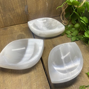 May include: Three white selenite leaf-shaped bowls. The bowls are smooth and have a slight sheen. They are arranged on a wooden surface, with green foliage in the background. The bowls are ideal for holding small items.