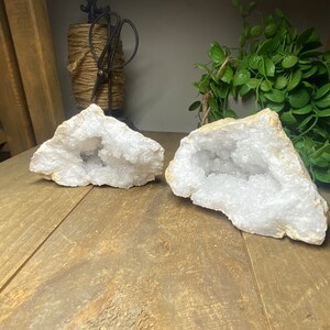 May include: Two white geode halves with crystal formations inside, displayed on a wooden surface. The geodes have a rough, textured exterior and a sparkling interior. The background includes a decorative spool of twine with scissors and greenery.