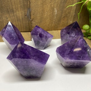 May include: Five amethyst crystal points with multiple facets. The crystals are a deep purple hue and have a polished, reflective surface. They are arranged on a white surface with a wooden background and some greenery.
