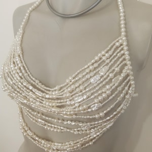 May include: A white pearl halter top with multiple strands of pearls cascading down. The top is displayed on a mannequin. The pearls vary in size and shape, creating a textured and elegant look. The top is designed to be worn as a garment.