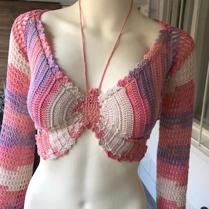 May include: A handmade, long-sleeved crochet top in a variety of pink, purple, and cream colors. The top features a plunging neckline and a delicate, scalloped edge. A thin, pink cord connects the two halves of the top.