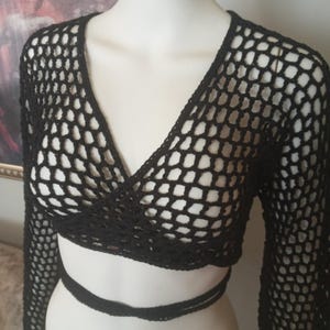May include: Black, open-weave, long-sleeved crop top with a V-neck design. The top features a wrap-around tie at the waist. The garment is displayed on a mannequin. The open weave design is visible throughout the top.
