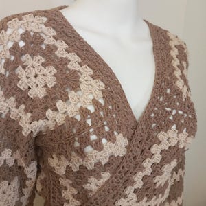 May include: A brown and beige crochet wrap top with long sleeves. The wrap top features a V-neck design and a geometric pattern. The garment is displayed on a mannequin.