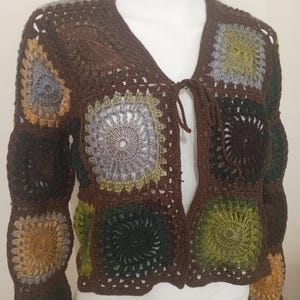 May include: A handmade crochet jacket in shades of brown, green, and gray. The jacket features a V-neck and long sleeves, with a tie closure at the neckline. The design incorporates square, circular motifs, creating a patchwork effect. The garment is displayed on a mannequin.