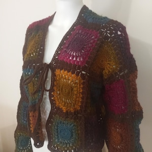 May include: A handmade crochet jacket with a patchwork design. The squares are in shades of brown, blue, orange, and purple. The jacket has long sleeves and a tie closure at the front. The open weave design adds a unique texture.