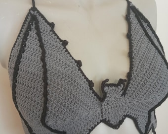 Puede incluir: Top de ganchillo gris con ribete negro y tirantes halter. El top presenta un diseño asimétrico inusual con un borde festoneado y pequeños detalles decorativos. La prenda hecha a mano se exhibe en un maniquí.