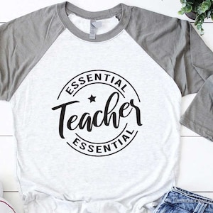 May include: A white and gray raglan t-shirt with a black circle graphic that says "Essential Teacher Essential".