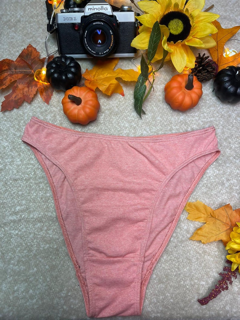 May include: A peach-colored cotton thong underwear with a high leg cut. The underwear is photographed on a white surface with fall-themed decorations.