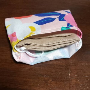 May include: A pink fabric pouch with a floral pattern and a beige fabric mask folded inside.