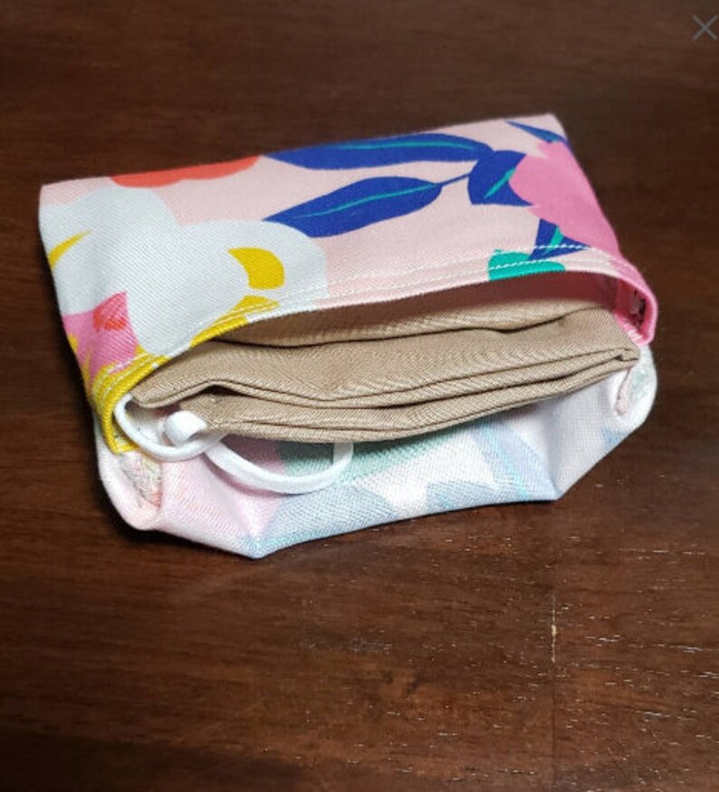 May include: A pink fabric pouch with a floral pattern and a beige fabric mask folded inside.