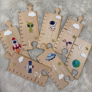May include: A set of six light-colored wooden puzzle pieces shaped like rulers, each featuring space-themed graphics including an alien, astronaut, rocket, satellite, UFO, telescope, and Earth, along with numbers one through six and white clouds.