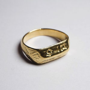 May include: A gold-colored ring with a flat, rectangular face featuring embossed lettering that appears to read S I R. The band has a textured, hammered finish with a geometric cross-hatch pattern engraved on the side of the ring shank.