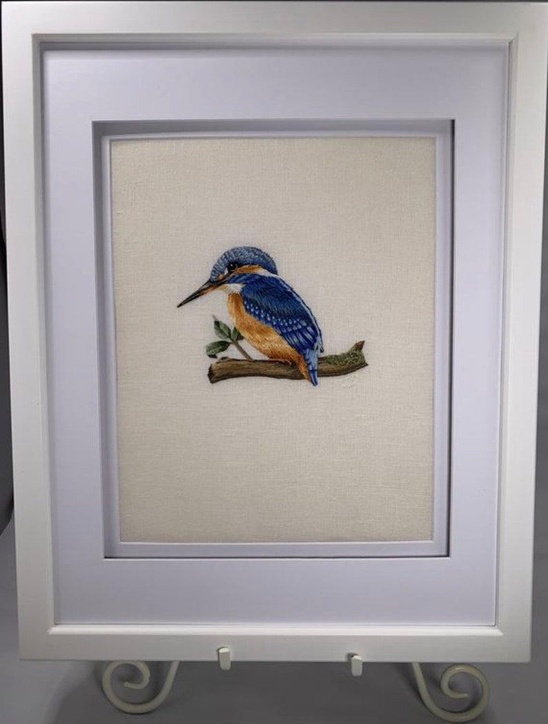 May include: A framed embroidery depicting a blue and orange kingfisher perched on a branch with green leaves.