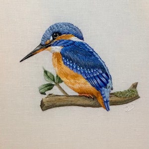 May include: A blue and orange kingfisher bird perched on a branch with green leaves. The bird is embroidered on a white fabric.