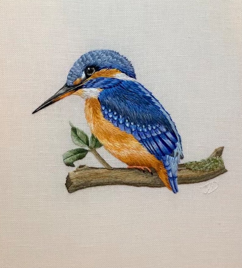 May include: A blue and orange kingfisher bird perched on a branch with green leaves. The bird is embroidered on a white fabric.