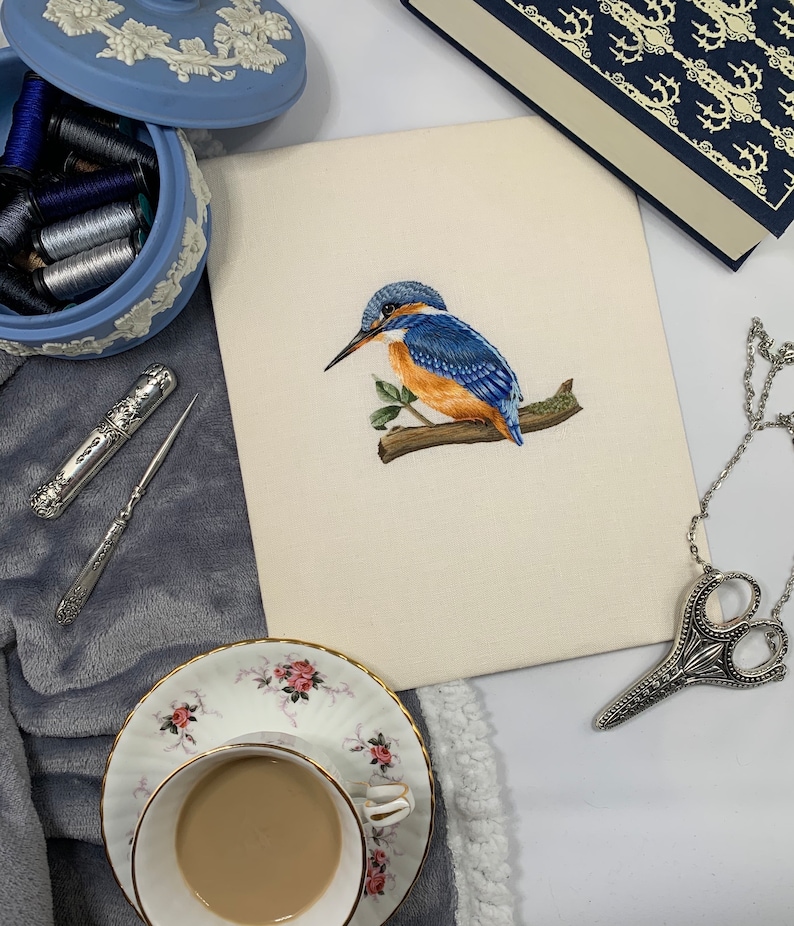 May include: A close-up of a needlepoint embroidery depicting a blue and orange kingfisher perched on a branch. The embroidery is on a white canvas and is surrounded by a white mount. The embroidery is on a table with a blue and white porcelain box, a cup of coffee, and a pair of silver scissors.