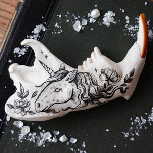 Puede incluir: Una mandíbula animal de color blanco hueso con una detallada ilustración en tinta negra de una cabeza de unicornio rodeada de motivos florales. El hueso tiene una textura lisa y un diente con punta marrón en la parte posterior, con estrellas.