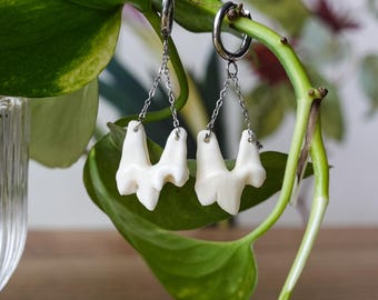 Puede incluir: Un par de pendientes de aro plateados con colgantes irregulares de color blanco roto en forma de diente, suspendidos por delicadas cadenas de metal plateado. Los colgantes tienen un acabado mate liso y una estructura orgánica ramificada.