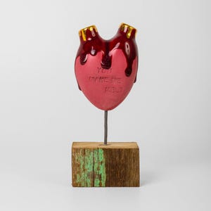 May include: A red heart-shaped sculpture with dripping red accents and gold-colored details. The text "YOU MAKE ME MELT" is inscribed on the front. The heart is mounted on a metal rod and a rustic wooden base.