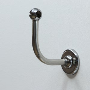Puede incluir: Un gancho de metal cromado con un extremo redondeado y una base circular. El gancho está montado en una pared blanca.
