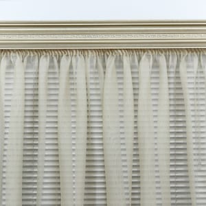 May include: A window treatment featuring a decorative, light-colored wooden valance above sheer, cream-colored curtains. The curtains hang in soft folds, partially obscuring the horizontal blinds behind them. The valance has a detailed, carved design.