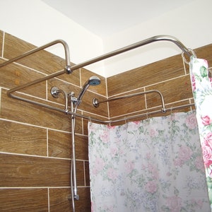 May include: A chrome shower curtain rail with two tiers, mounted in a corner of a bathroom with brown tiled walls. The rail is holding a white shower curtain with a floral pattern.