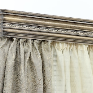 May include: A decorative wooden cornice in shades of brown and gold, mounted above a window. The cornice has detailed moulding and a decorative border. Below, gathered curtains in beige and cream tones add a soft contrast.