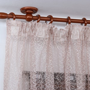 Può includere: Un'asta per tende in legno marrone con un terminale decorativo e anelli per appendere le tende. Le tende sono di colore marrone chiaro e trasparenti, con un motivo delicato.