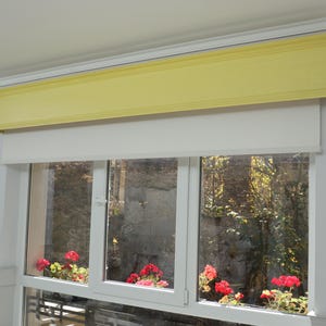 May include: A window featuring a white frame, white blinds, and a yellow valance. Red geraniums in window boxes provide colour. The window offers a view of a garden.