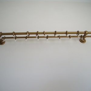 Könnte beinhalten: Eine goldene Doppel-Gardinenstange aus Metall mit Endkappen an beiden Enden. Die Stange ist an einer weißen Wand montiert und hat weiße Kunststoff-Gardinenringe.
