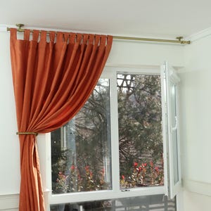 Könnte beinhalten: Orangefarbene Vorhänge mit einer Messinggardinenstange, die an der Decke befestigt ist. Die Vorhänge sind mit einem Messingvorhangbinder zurück gebunden. Hinter den Vorhängen befindet sich ein Fenster mit einem weißen Rahmen. Das Fenster ist geöffnet und zeigt einen Blick auf Bäume im Freien.