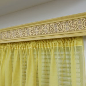 May include: A yellow sheer curtain with a decorative wooden valance featuring a carved design. The valance is painted a light brown color.