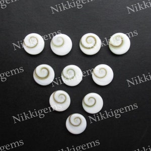 May include: Ten white, circular gemstones with a spiral pattern at their centre. The spiral is a light brown colour. The gemstones are displayed on a black surface. The image is watermarked with the text "Nikkigems".