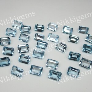 May include: A collection of 20 light blue topaz gemstones in an emerald cut. The gemstones are faceted and have a clear, glassy appearance.