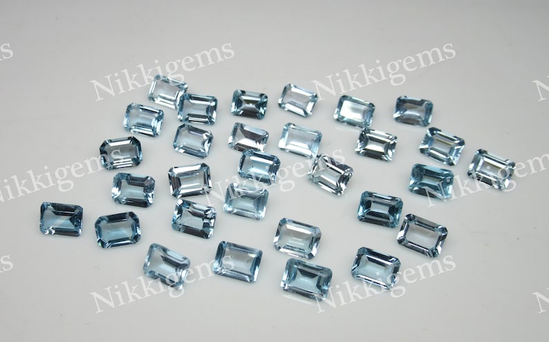 May include: A collection of 20 light blue topaz gemstones in an emerald cut. The gemstones are faceted and have a clear, glassy appearance.