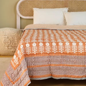 Op de afbeelding: Een oranje quilt met een wit bloemenpatroon en een bruine zigzagrand. Het bed heeft witte kussens en een lichtroze hoofdbord met een geweven rotan achterkant. Afmetingen: ongeveer 200 cm x 200 cm.