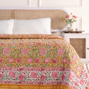 Könnte beinhalten: Eine gelbe Blumensteppdecke mit rosa Blüten und grünen Blättern. Die Steppdecke hat einen weißen Rand mit rosa Blumendesigns und eine gelbe Bordüre. Die Steppdecke liegt auf einem Bett mit weißen Kissen und einem gewebten Kopfteil. Das Gesamtthema ist Wohnkultur.