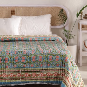 May include: A floral patterned quilt in shades of blue, coral, and green, draped over a bed. The quilt features a repeating floral design and a striped border. White pillows with pom-pom trim rest on the bed.