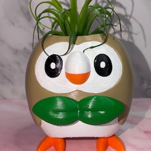 Peut inclure: Un pot de fleurs rond de couleur beige en forme de hibou avec un visage blanc, des yeux noirs, un bec orange et un nœud papillon en forme de feuille verte. Le pot possède des pieds orange vif et contient une petite plante verte.