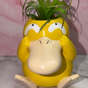May include: A yellow duck-shaped planter with a large beige beak, white eyes, and two hands held to its head. The planter features a green air plant sprouting from the top opening, creating a bright and colorful decorative container for greenery.