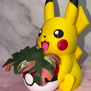 May include: A yellow Pikachu figurine with black-tipped ears and red cheeks, holding a red, white, and black Poké Ball planter. The planter contains a small artificial plant with green and pink variegated leaves, featuring a visible 3D-printed texture.