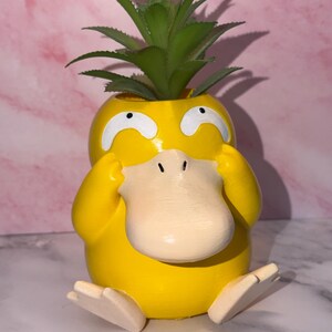 May include: A yellow, duck-like planter with a large, flat, beige beak and white eyes, featuring a green, spiky succulent plant emerging from the top. The figure is in a seated position with its hands held up to its head and beige feet extended.