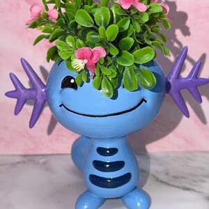 May include: A light blue axolotl-shaped planter with purple external gills, a smiling face, and dark blue horizontal stripes on the torso. The planter is filled with green foliage and small pink flowers, creating a decorative container for plants.