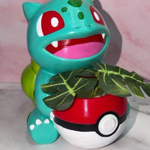 May include: A teal creature figurine with red eyes and a pink mouth holding a red, white, and black spherical planter. The planter contains a small artificial green plant with dark leaves featuring light green veins, creating a decorative display.