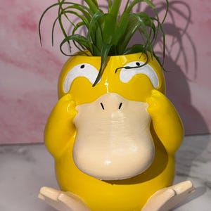 May include: A yellow planter shaped like the character Psyduck with a beige bill and feet, white eyes, and hands held to its head. A green air plant with thin, spiky leaves is placed inside the top opening of the yellow creature-shaped container.