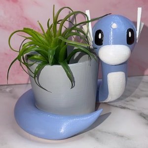 May include: A light blue, serpentine-shaped planter featuring a smooth, rounded head with large black eyes, a white snout, and white fin-like ears. A small, grey, cylindrical pot is nestled within the body, containing a green air plant.