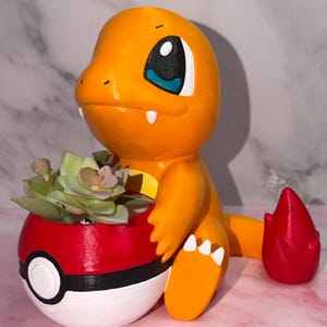 May include: A 3D printed orange Charmander figure holding a red, white, and black Poké Ball planter. The planter contains a small green succulent plant. The figure has a bright red flame at the tip of its tail and white claws on its feet.