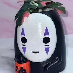 May include: A black and white No-Face planter with a small orange lantern. The planter has a white face with black eyes and purple accents. Green and pink leaves sprout from the top. The lantern has Japanese characters.